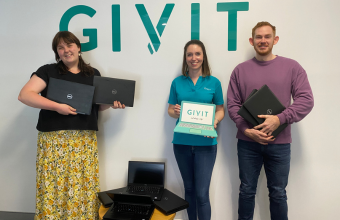 GIVIT staff member Danni and volunteer Hayden with a staff member from Queenslanders with Disability Network. All are holding laptops and standing in front of the GIVIT logo.