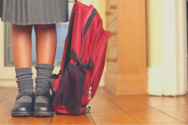 A young child wearing black school shoes, grey socks and holding a red backpack on the floor